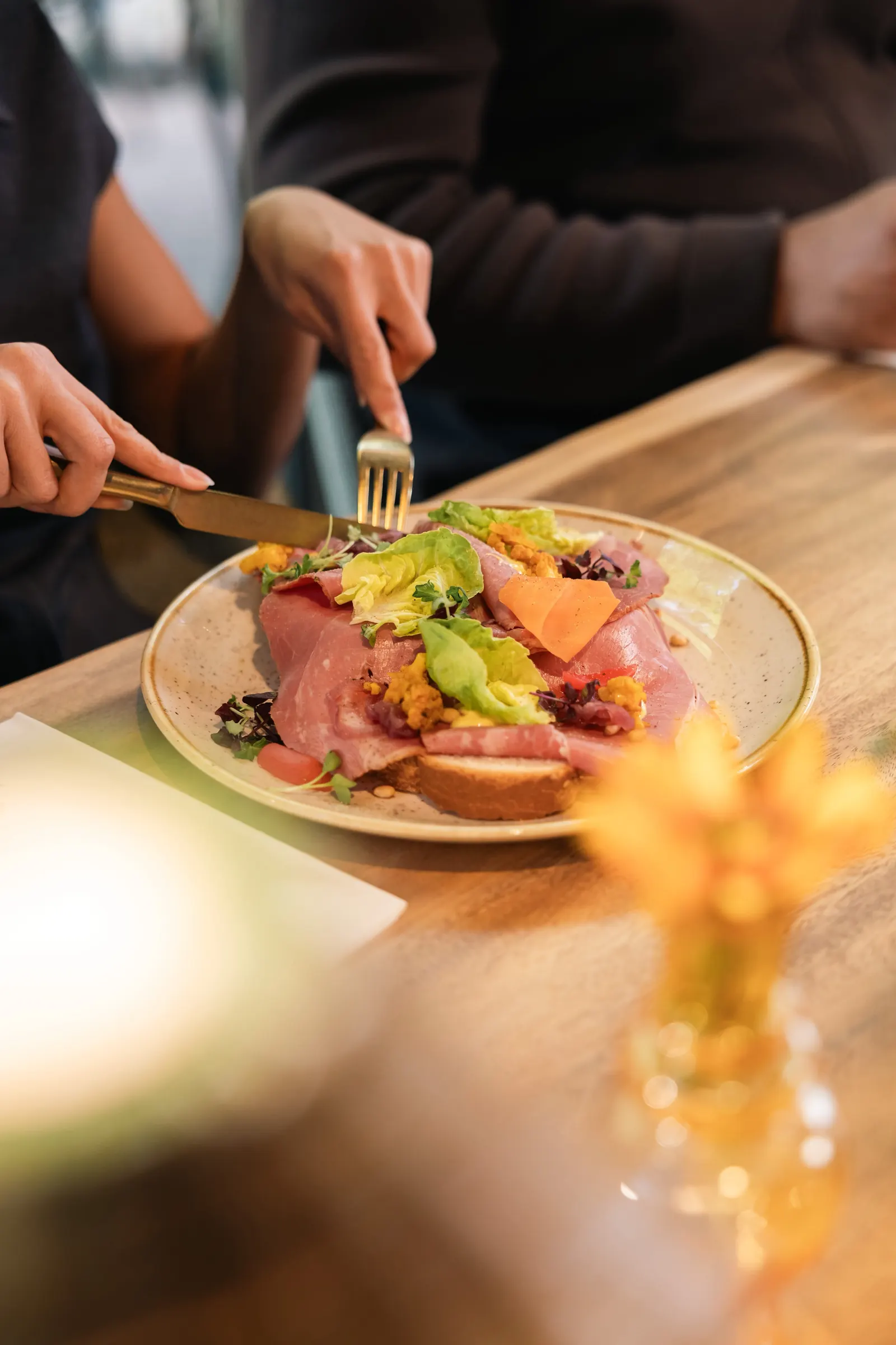 Een gast snijdt in een broodje met dun gesneden rood vlees, sla, een schijfje wortel en kiemgroente, op een gespikkeld bord op een houten tafel.