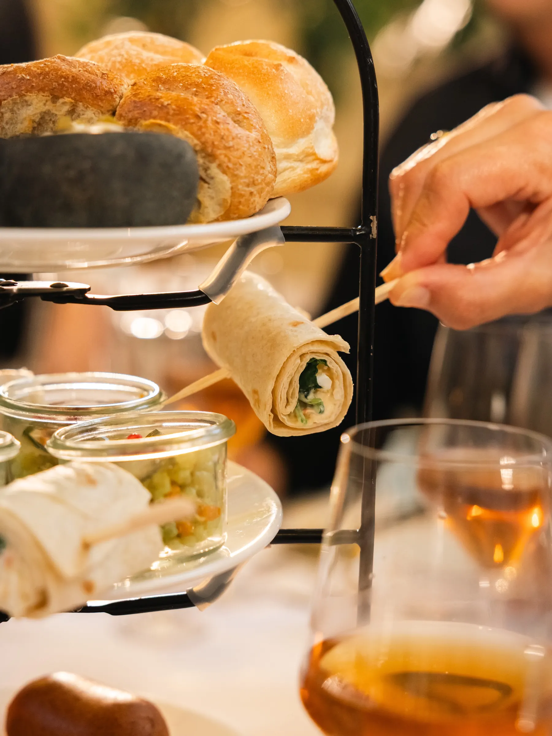 Etagère van de high tea bij Jacob van Bijstervelt van dichtbij: broodjes bovenin, wraps aan een prikker en glaasjes met groentesalade, en een hand die een wrap pakt.