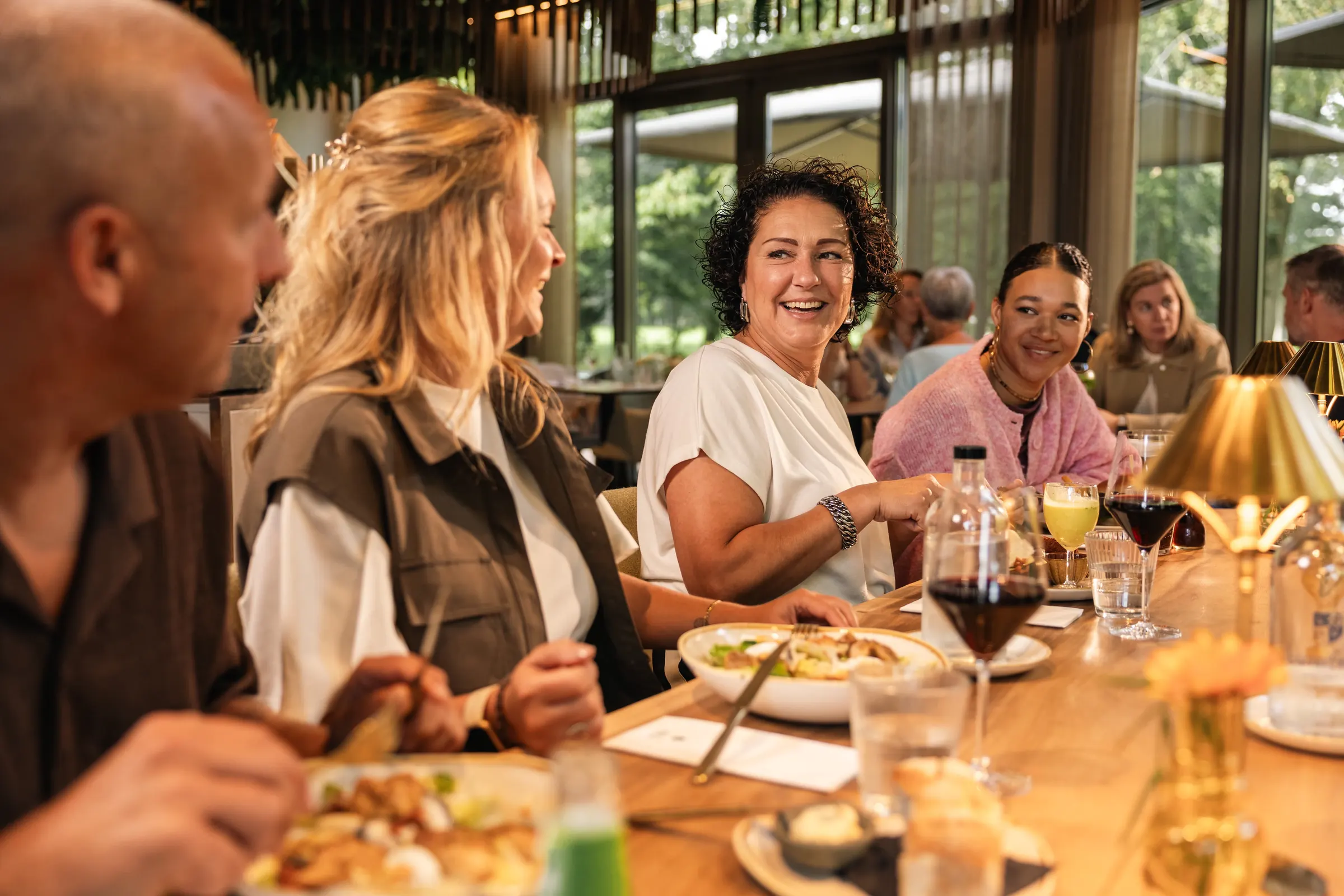 Gasten lachen en eten samen aan een lange houten tafel in Brasserie Jacob van Bijstervelt, met rode wijn, gouden tafellampjes en grote ramen naar het groen.
