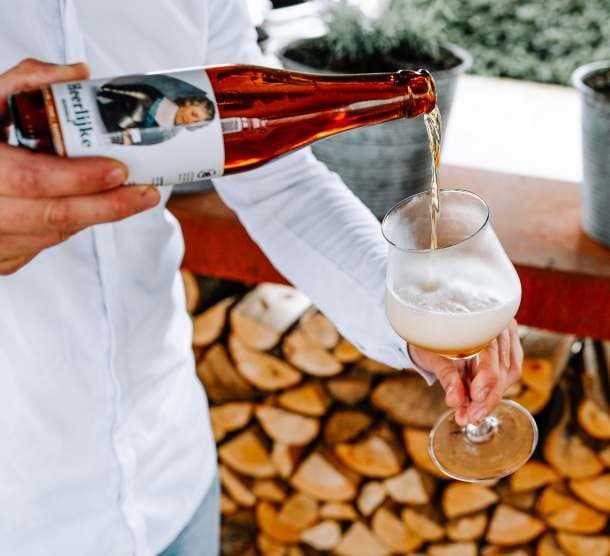 Een medewerker schenkt een fles Heerlijke Hendrik uit in een bierglas, voor een stapel openhaardhout.