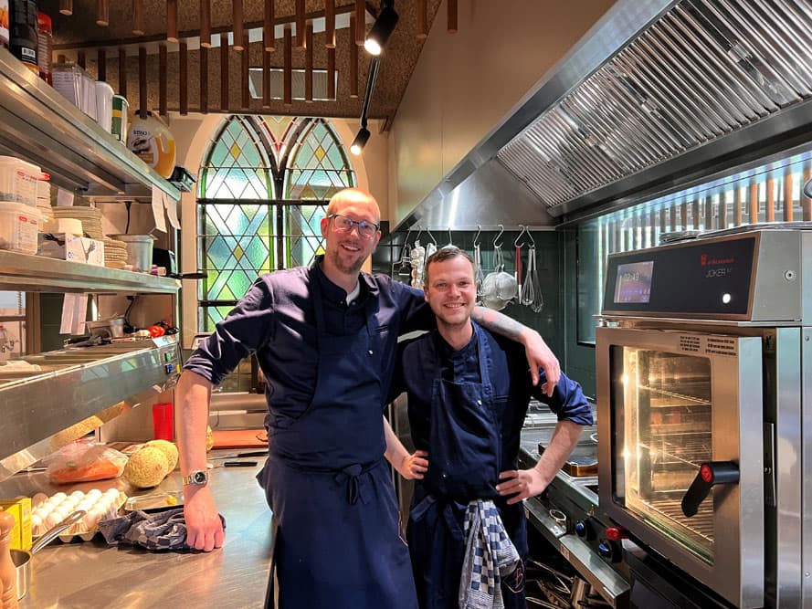Twee koks in donkerblauwe schorten lachen in de keuken, met een gotisch glas-in-loodraam achter hen.