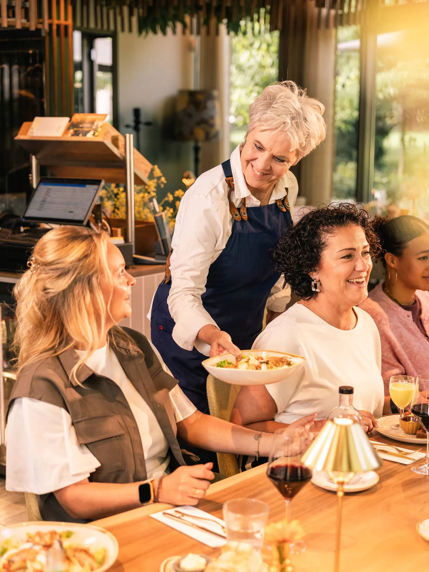 Een medewerker in een blauw schort zet lachend een lunchgerecht op tafel bij een groep gasten in Brasserie Jacob van Bijstervelt, met de tuin achter de ramen.