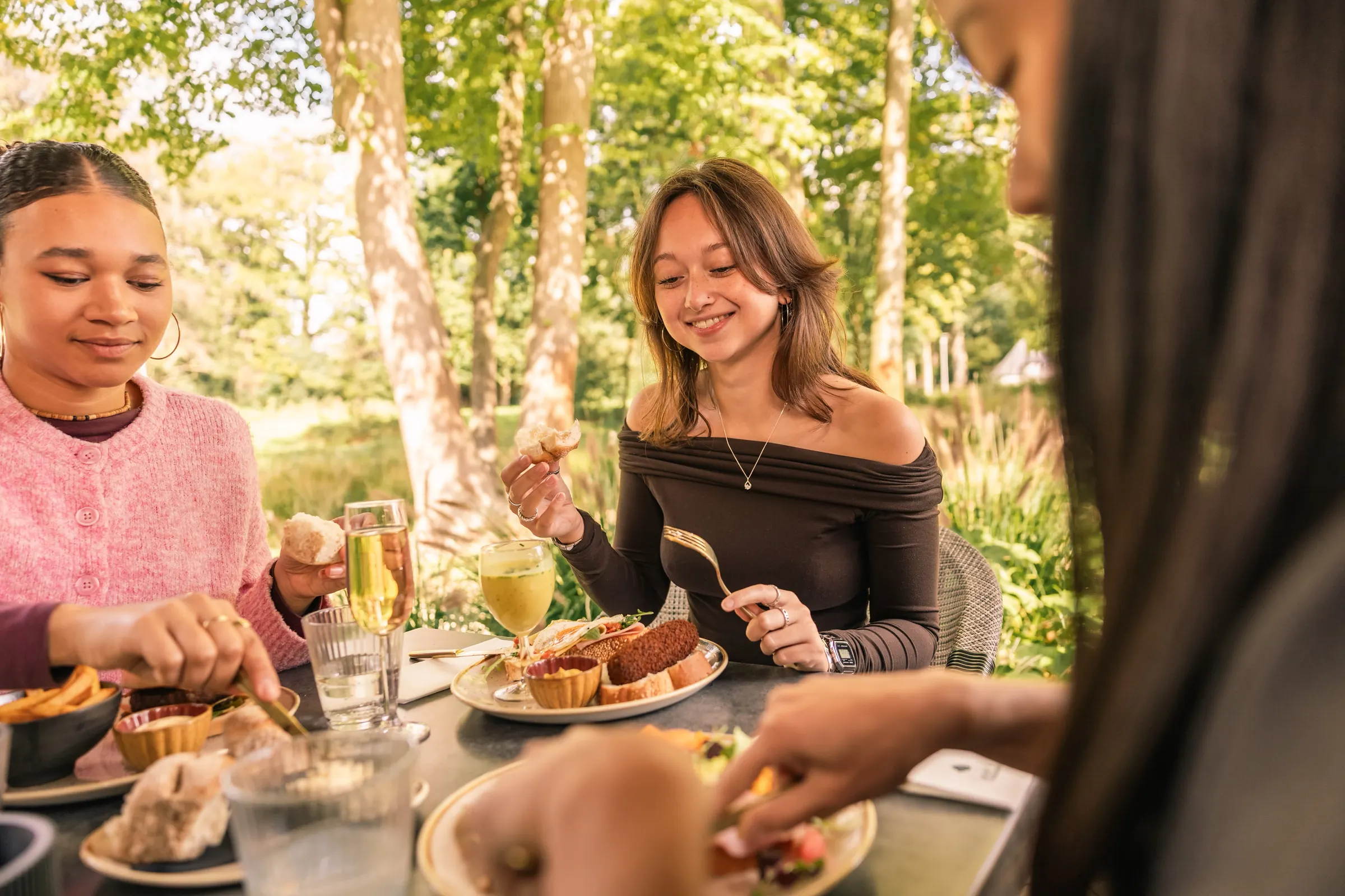 Drie vriendinnen eten samen aan een tafel in de tuin, onder hoge bomen in de zon, met hapjes, broodjes en glazen op tafel.