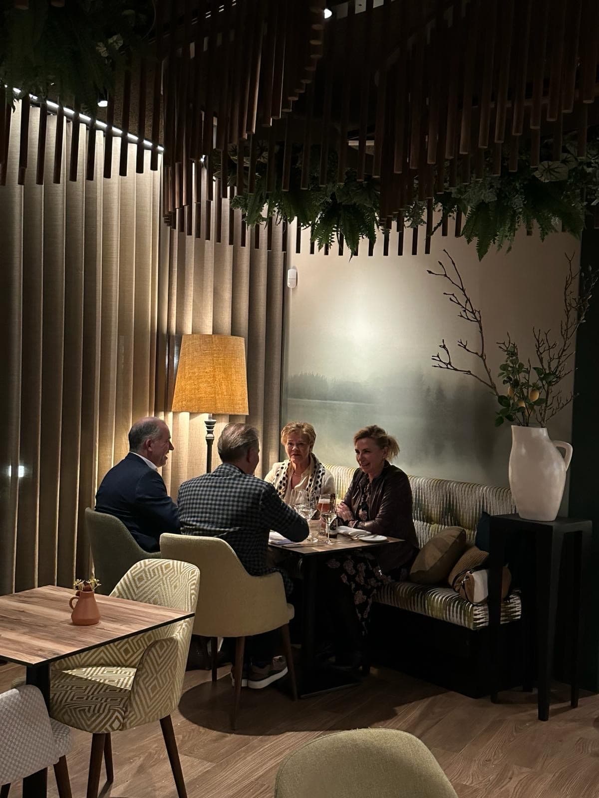 Vier gasten in gesprek aan een tafel in de brasserie, in het warme licht van een staande lamp.