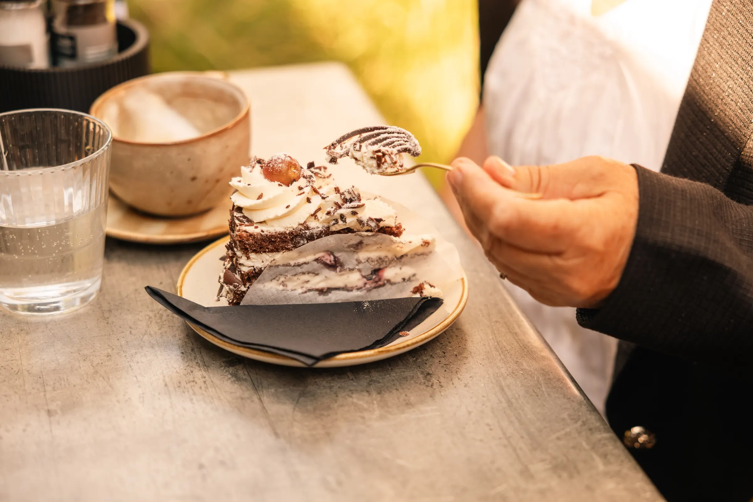 Een hand prikt met een vorkje in een punt chocoladetaart met slagroom en een kers, naast een cappuccino en een glas water.