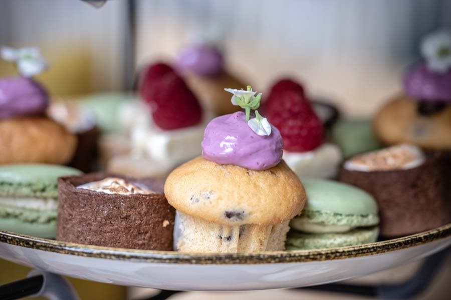 Zoete lekkernijen van de high tea: een muffin met paarse room en een eetbaar bloemetje, frambozen, macarons en chocoladetaartjes.